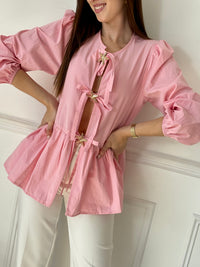 Cette blouse rose Adalia allie élégance et confort avec ses manches longues bouffantes, son col rond et ses poignets élastiques. Les liens à nouer sur le devant et les volants délicats sur le bas apportent une touche féminine et raffinée, idéale pour sublimer votre style au quotidien.