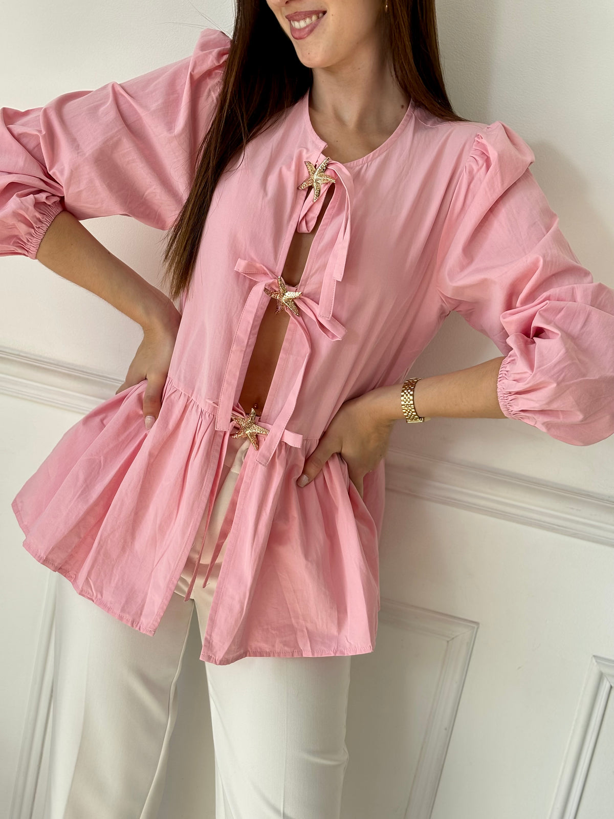 Cette blouse rose Adalia allie élégance et confort avec ses manches longues bouffantes, son col rond et ses poignets élastiques. Les liens à nouer sur le devant et les volants délicats sur le bas apportent une touche féminine et raffinée, idéale pour sublimer votre style au quotidien.