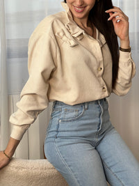Chemise beige en velours côtelé pour femme, dotée de manches longues et d'un col rond orné de volants. Nœuds latéraux et boutons discrets sur toute la longueur et aux poignets pour un style raffiné et élégant.