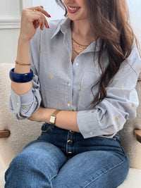 La chemise Sélène en coton combine style intemporel et détails délicats. Rayures verticales bleu et écru, cœurs dorés brodés et col chemise structuré créent une allure sophistiquée. Manches longues avec boutons aux poignets, poche poitrine et fermeture boutonnée complète.