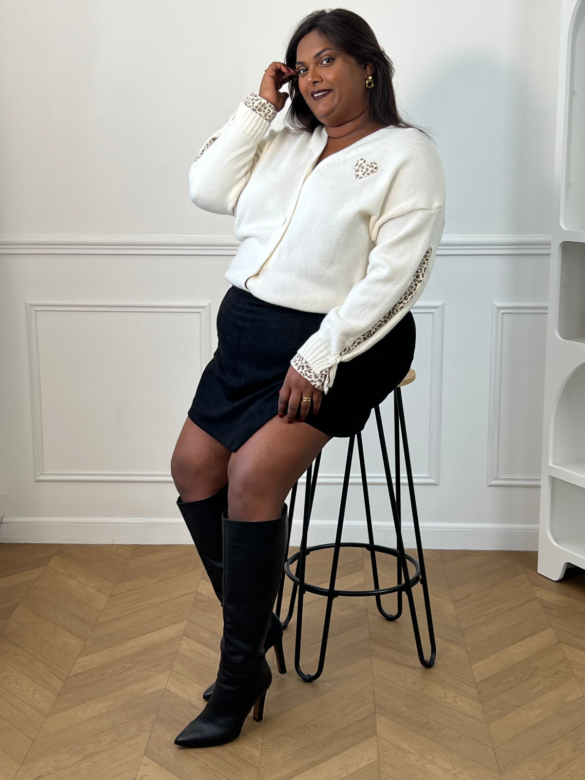 Gilet beige Stella porté en position assise sur un tabouret, avec une jupe-short noire et des bottes noires.