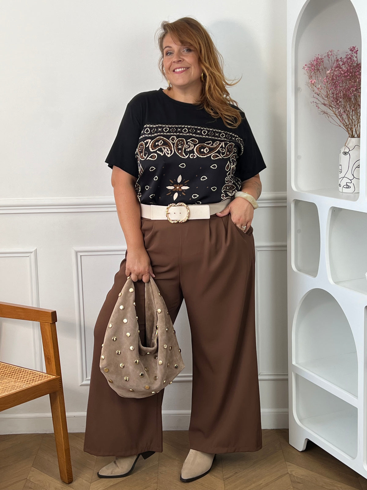 Tee-shirt noir Livana grande taille porté de face avec un pantalon large marron, une ceinture écrue et un sac taupe à clous.