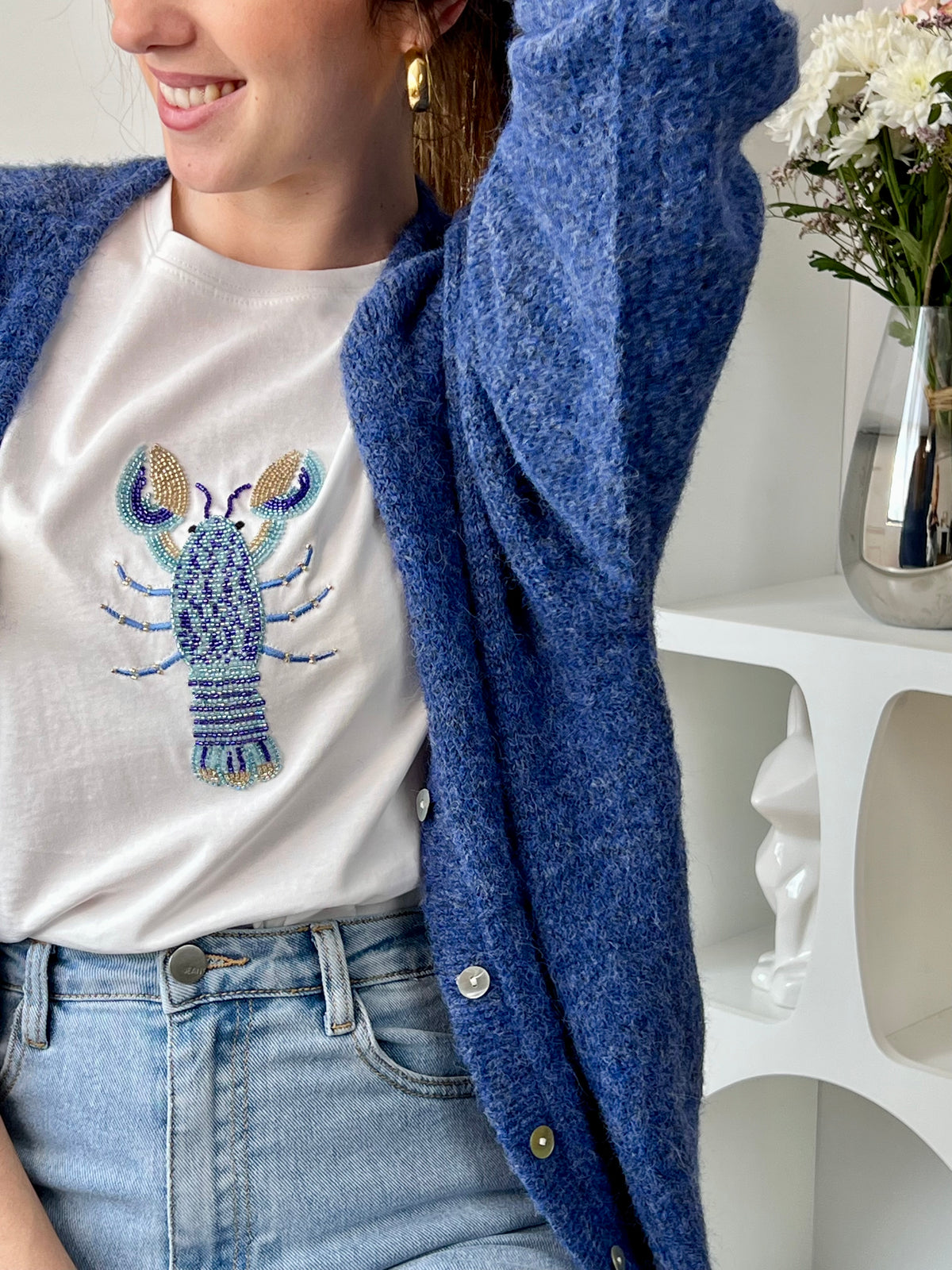 Tee-shirt écru femme en 100% coton, parfait pour un look décontracté et tendance. Avec ses manches courtes et son col rond, il offre un confort idéal au quotidien. Son motif homard en perles sur le devant apporte une touche originale et élégante. Facile à associer, ce tee-shirt aux détails écru, bleu et doré est idéal pour une tenue estivale chic et moderne.